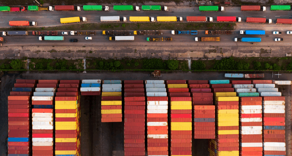 Luftaufnahme eines Hafens mit zahlreichen bunten Containern und Lkw-Aufliegern, die auf mehreren Spuren nebeneinander geparkt sind.